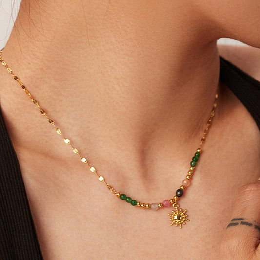 Gold necklace with green and red beads and a sun pendant on a person's neck.